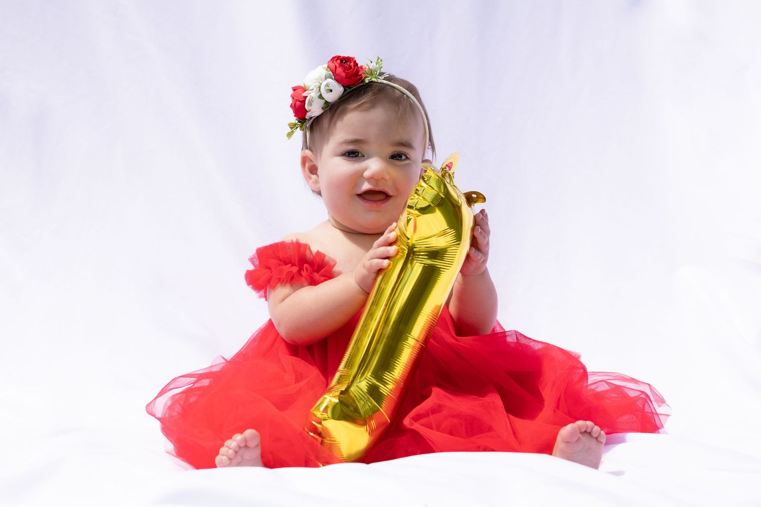 Baby in red tulle dress holding gold Christmas ornament against white background.
