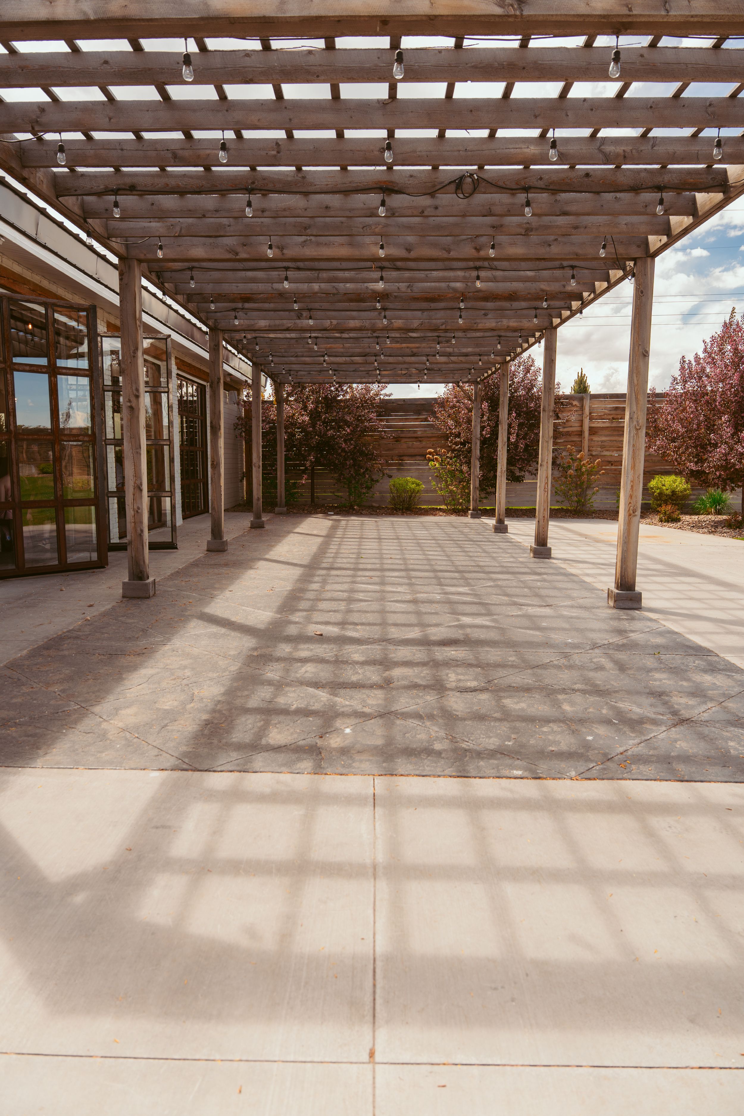 A covered walkway with wooden pergola beams casting geometric shadows on concrete path surrounded by landscaping.