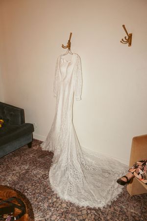 Elegant white lace wedding dress with long train hanging on wall hook in room with dark couch.