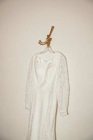 Elegant white lace wedding dress hanging on golden hanger against neutral wall.