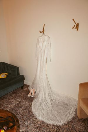 Full-length white lace wedding gown with train displayed on wall hooks with shoes below.