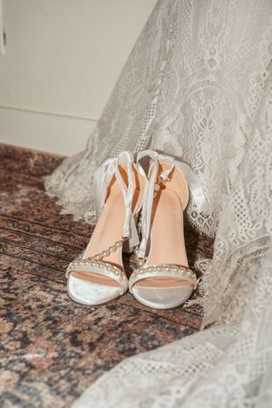 Elegant bridal sandals with delicate straps resting on a wooden floor next to lace wedding dress fabric.