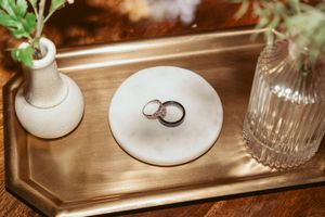 Wedding rings sit elegantly on a wooden surface near decorative white candle holders and glass bottles.