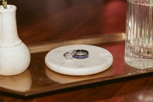 Two wedding rings rest on white ceramic holders alongside glass bottles on a polished wooden surface.
