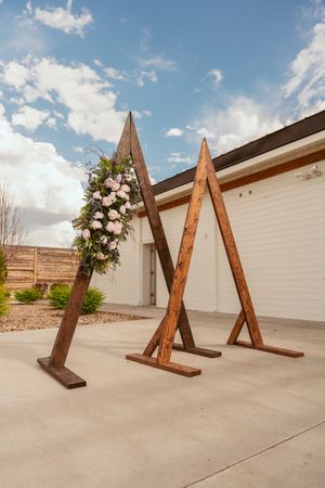 Modern wooden triangle wedding arch decorated with pink and white floral arrangement stands on concrete patio.