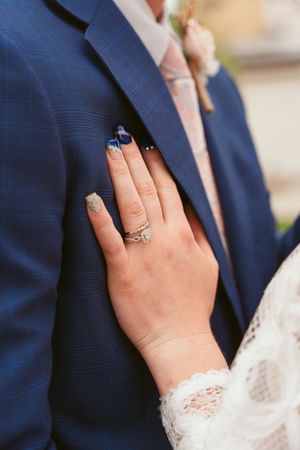 A hand wearing a delicate lace sleeve and sparkling engagement ring rests against a navy blue suit jacket.