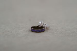 Wedding ring set featuring a diamond engagement ring and purple-accented band on a gray textured surface.