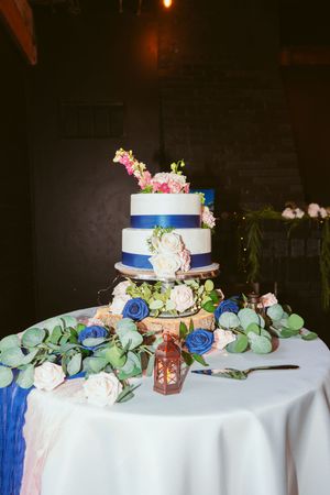 Three-tiered white wedding cake decorated with blue ribbons, pink and white flowers, and eucalyptus on a round table.