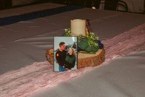 A rustic wood slice centerpiece with a white candle and photo frame decorated with greenery on a pink tablecloth.