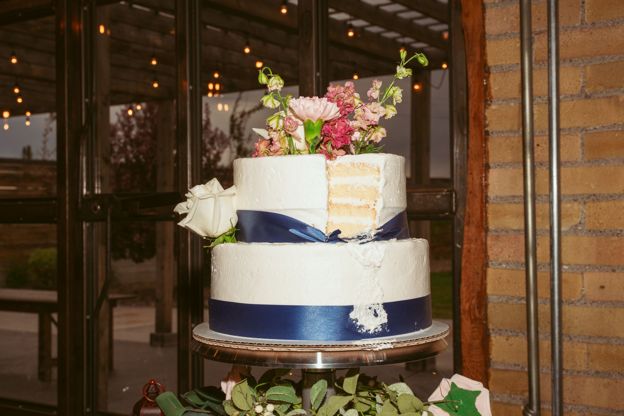 Two-tier wedding cake decorated with fresh flowers and navy blue ribbon on a rustic display.