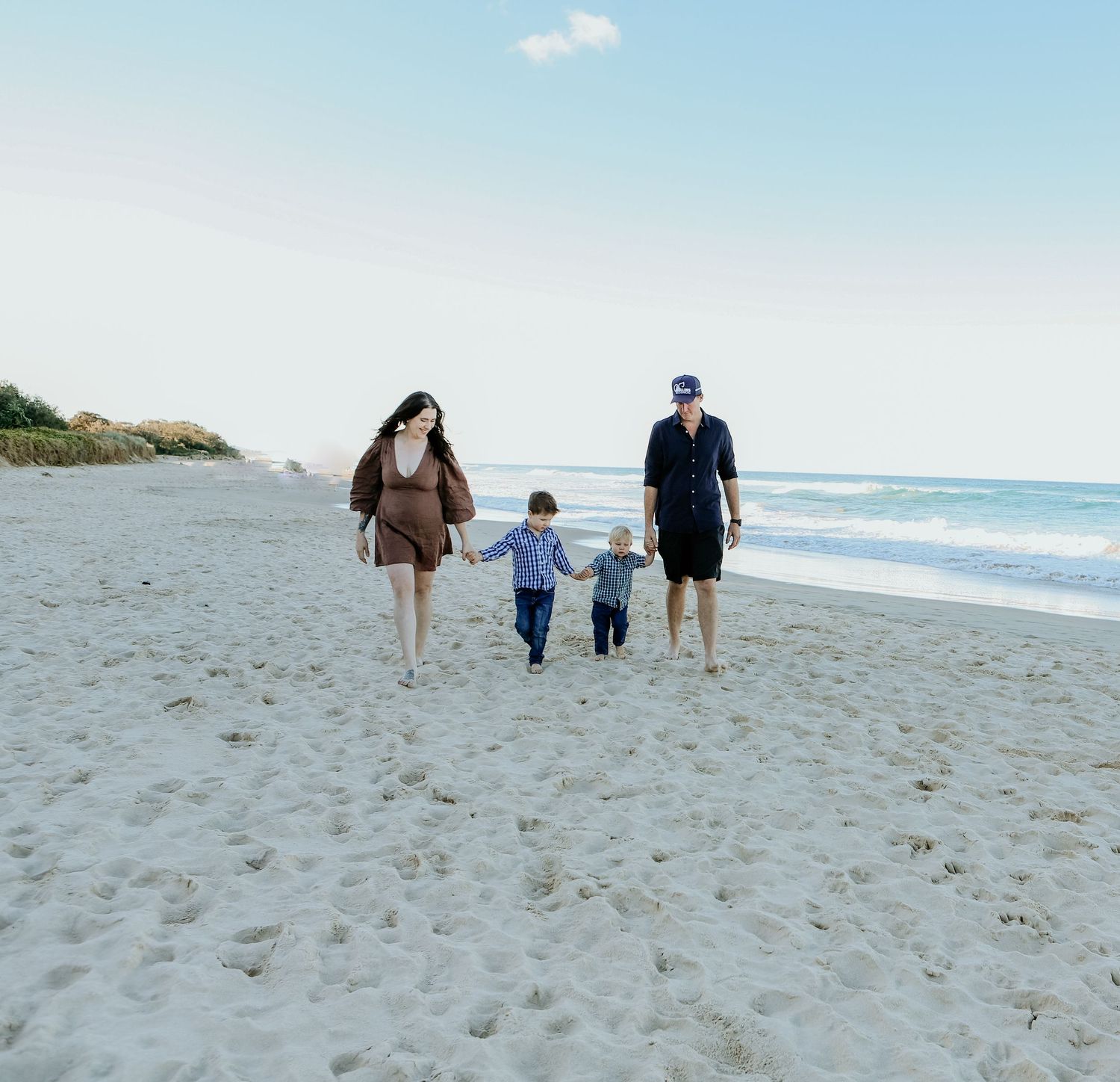 Three people walk hand in hand along a sunny beach shoreline with waves rolling in and blue skies above.
