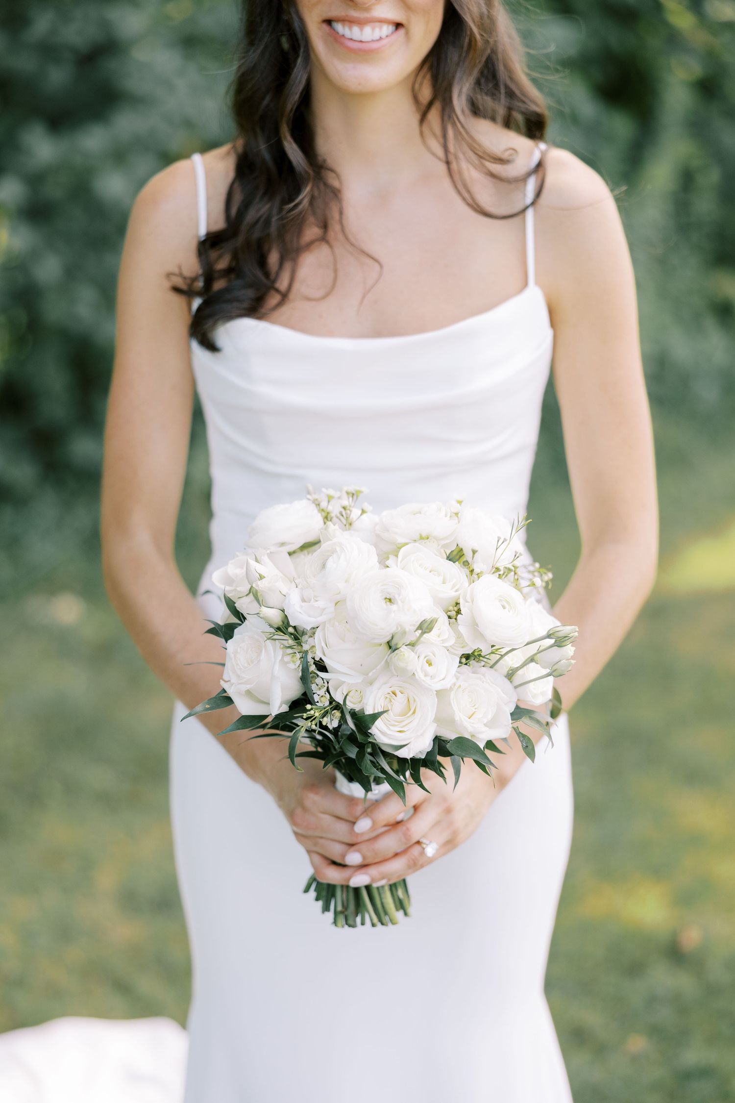 A bride in a sleek white spaghetti strap wedding dress holds an elegant bouquet of white peonies and delicate greenery.