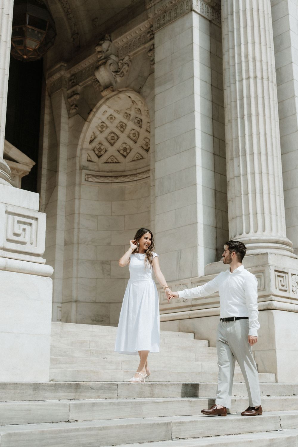A couple in white and light gray attire pose elegantly on marble steps of a grand neoclassical building.