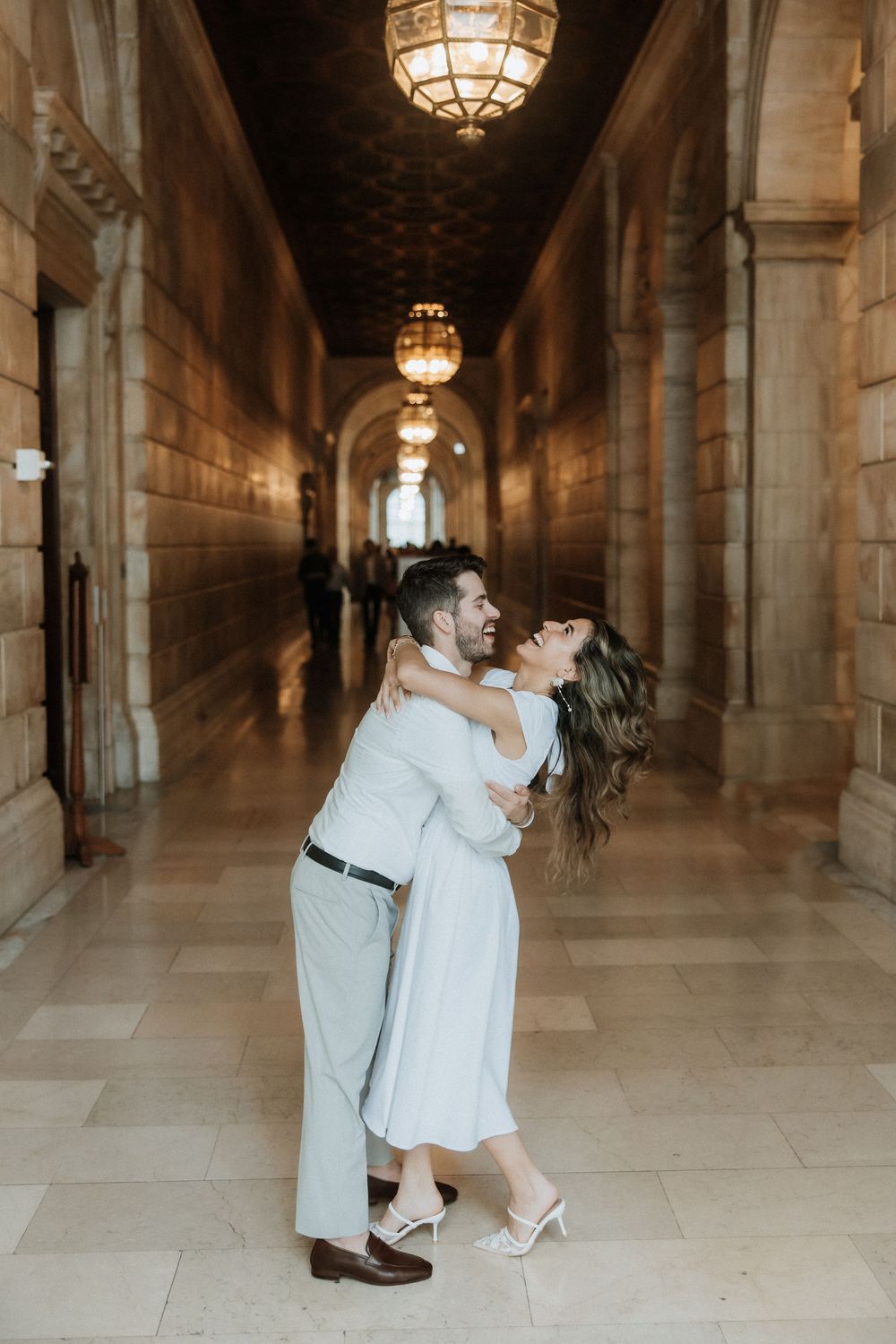 A couple dances romantically in an ornate hallway with stone columns and vintage lighting fixtures.