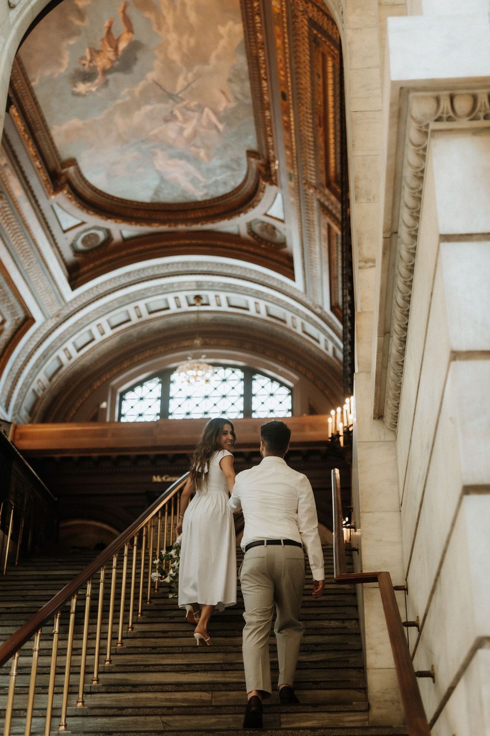Couple ascending grand marble staircase beneath ornate baroque ceiling with painted fresco in historic building.