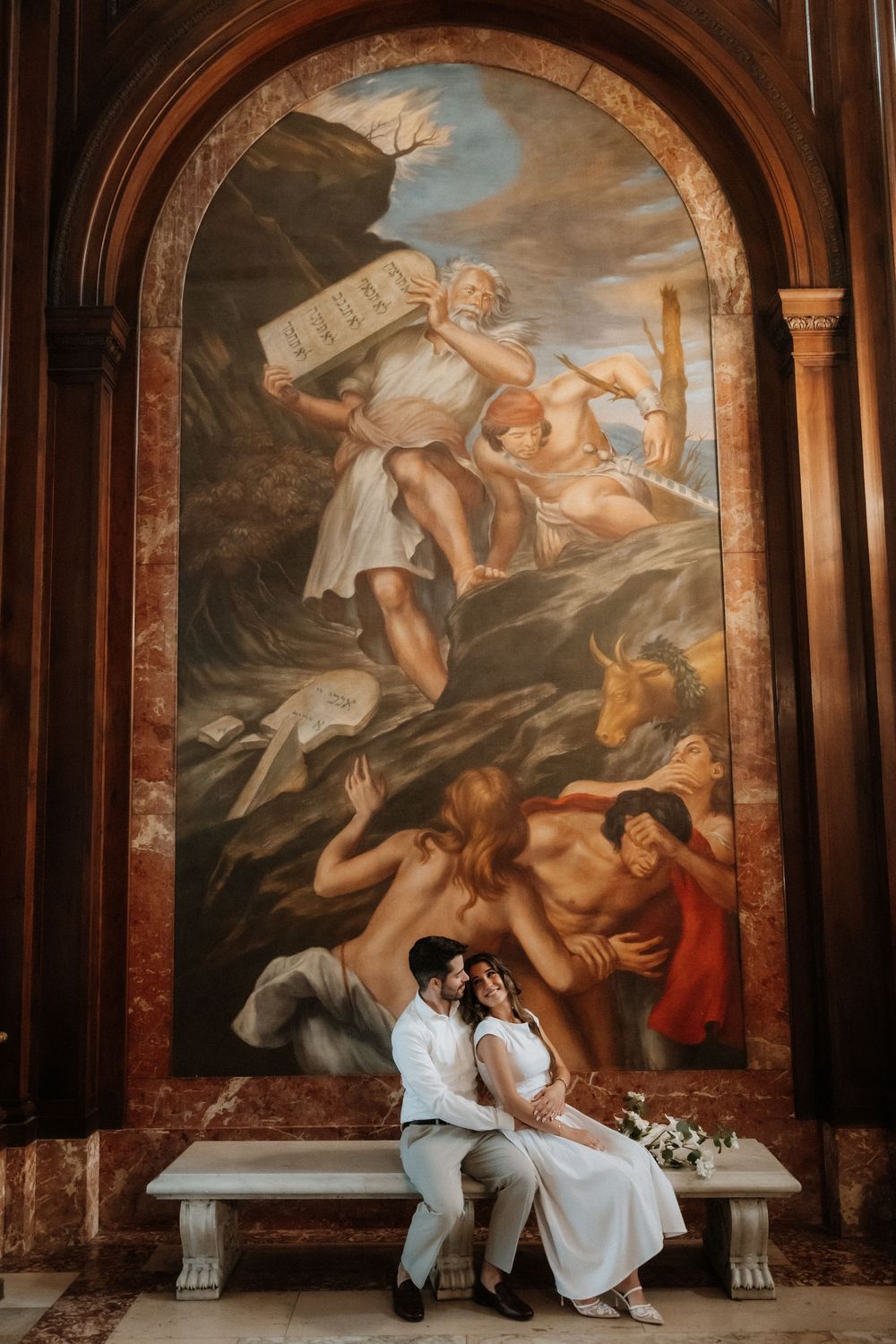 A couple in white outfits sits on a stone bench beneath grand Renaissance-style murals depicting mythological scenes.