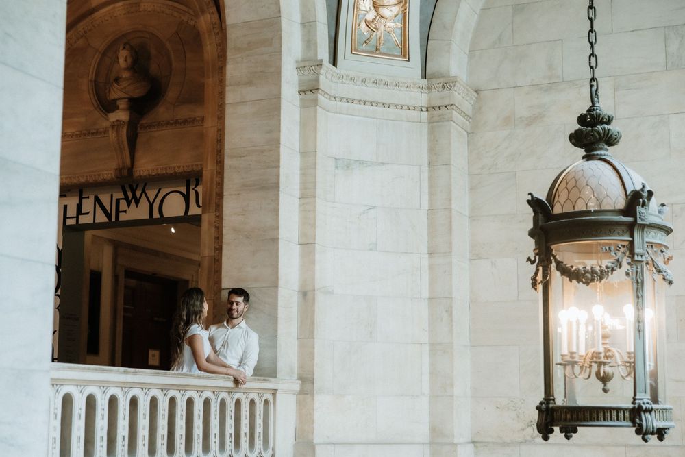 A couple stands on a white marble balcony with ornate architectural details and a hanging vintage lantern.