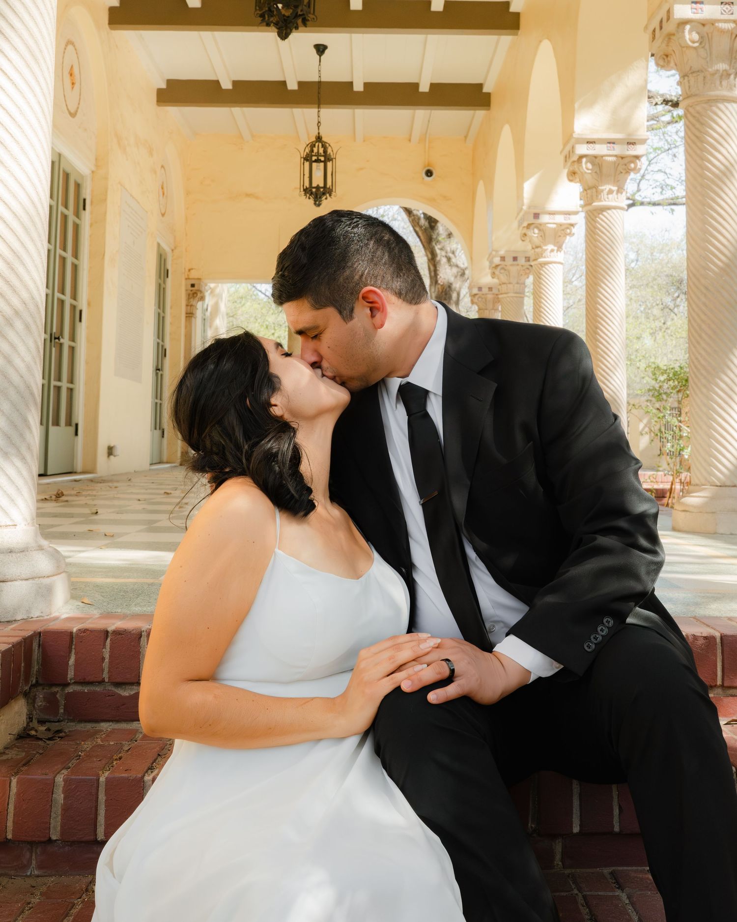 Couple shares tender moment on outdoor bench in warm sunlit colonnade.