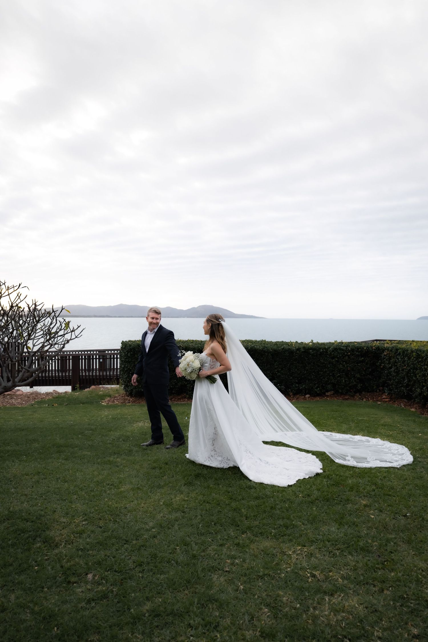 Wedding couple poses with flowing veil on scenic coastal grounds.