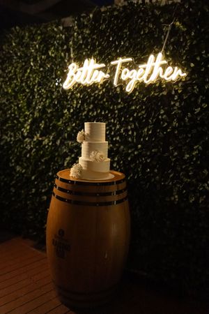 White wedding cake displayed on wooden barrel with illuminated 'Together' sign in background.