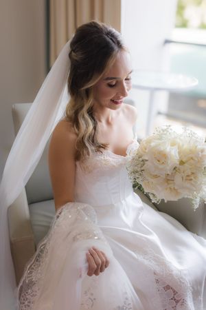 Bride in white lace dress sitting by window with cream colored bouquet.