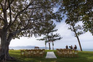 Outdoor wedding ceremony setup with wooden chairs under trees overlooking ocean view.