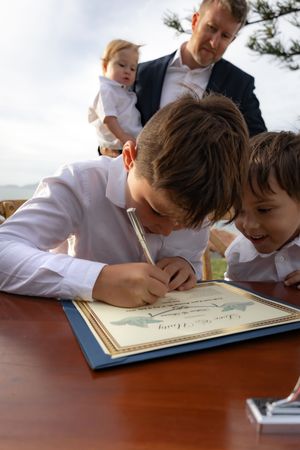 Children leaning over to sign wedding guest book on wooden table.