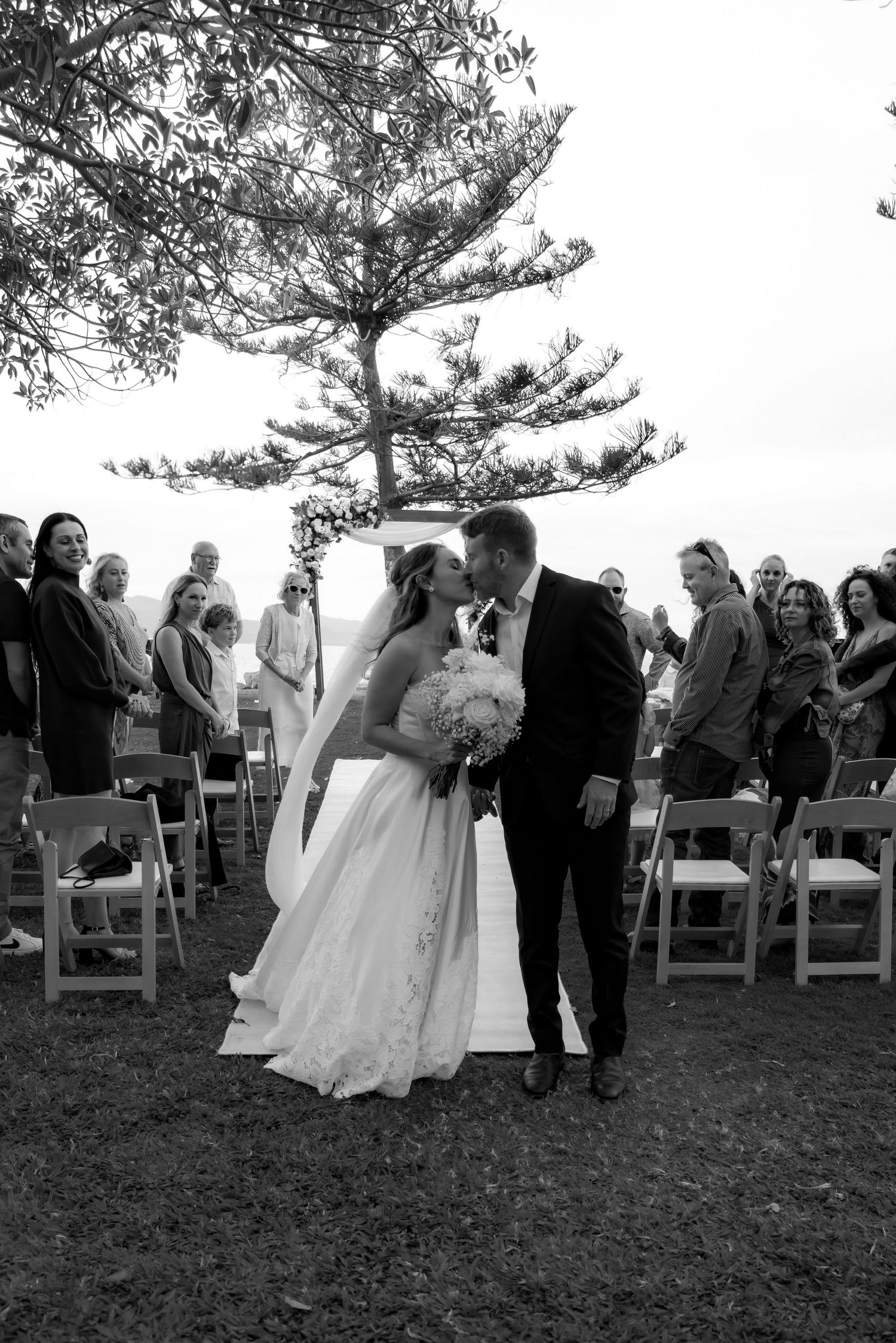 Wedding ceremony guests gather outdoors under pine trees for a celebration.