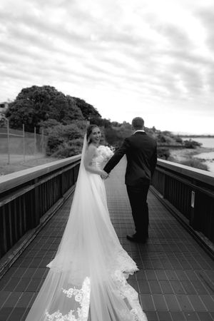 A couple walks along a wooden boardwalk bridge in formal wedding attire at sunset.