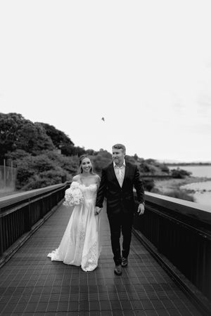 A bride in white dress and groom in black suit walk together on a wooden boardwalk.