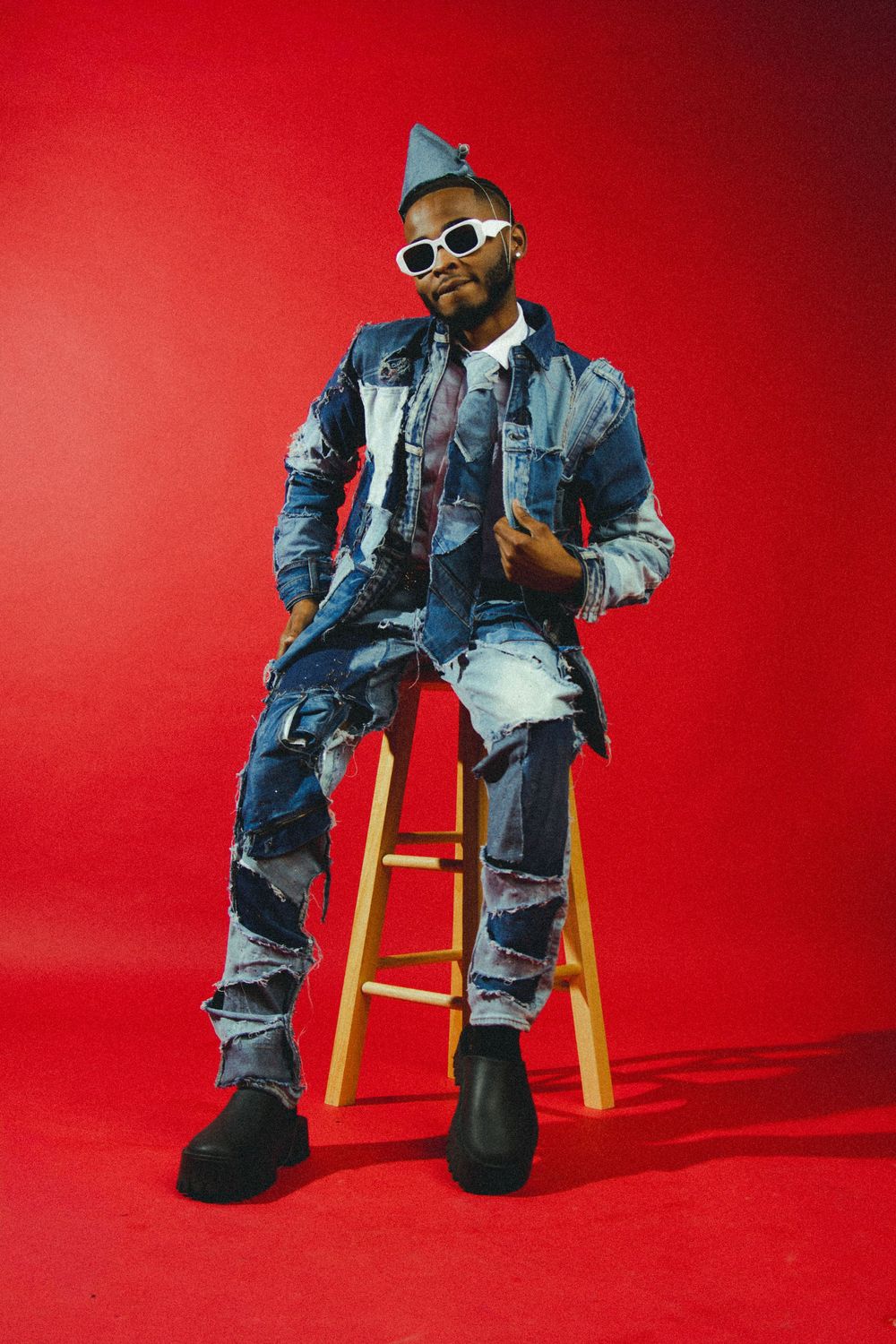 Person in patchwork denim ensemble poses on wooden stool with red studio backdrop.