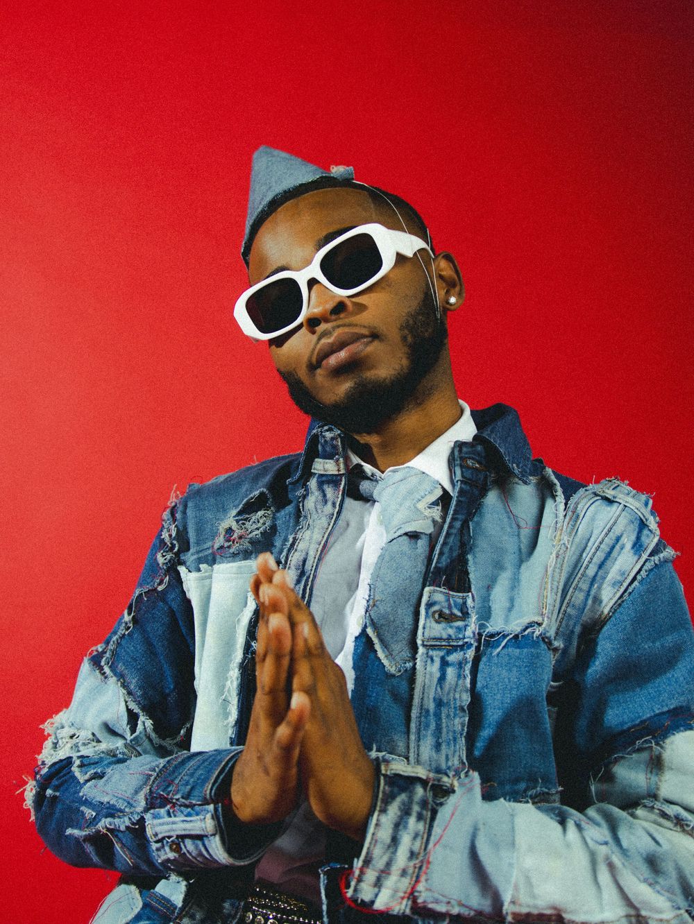 Person in distressed denim jacket and white sunglasses poses against vibrant red backdrop.