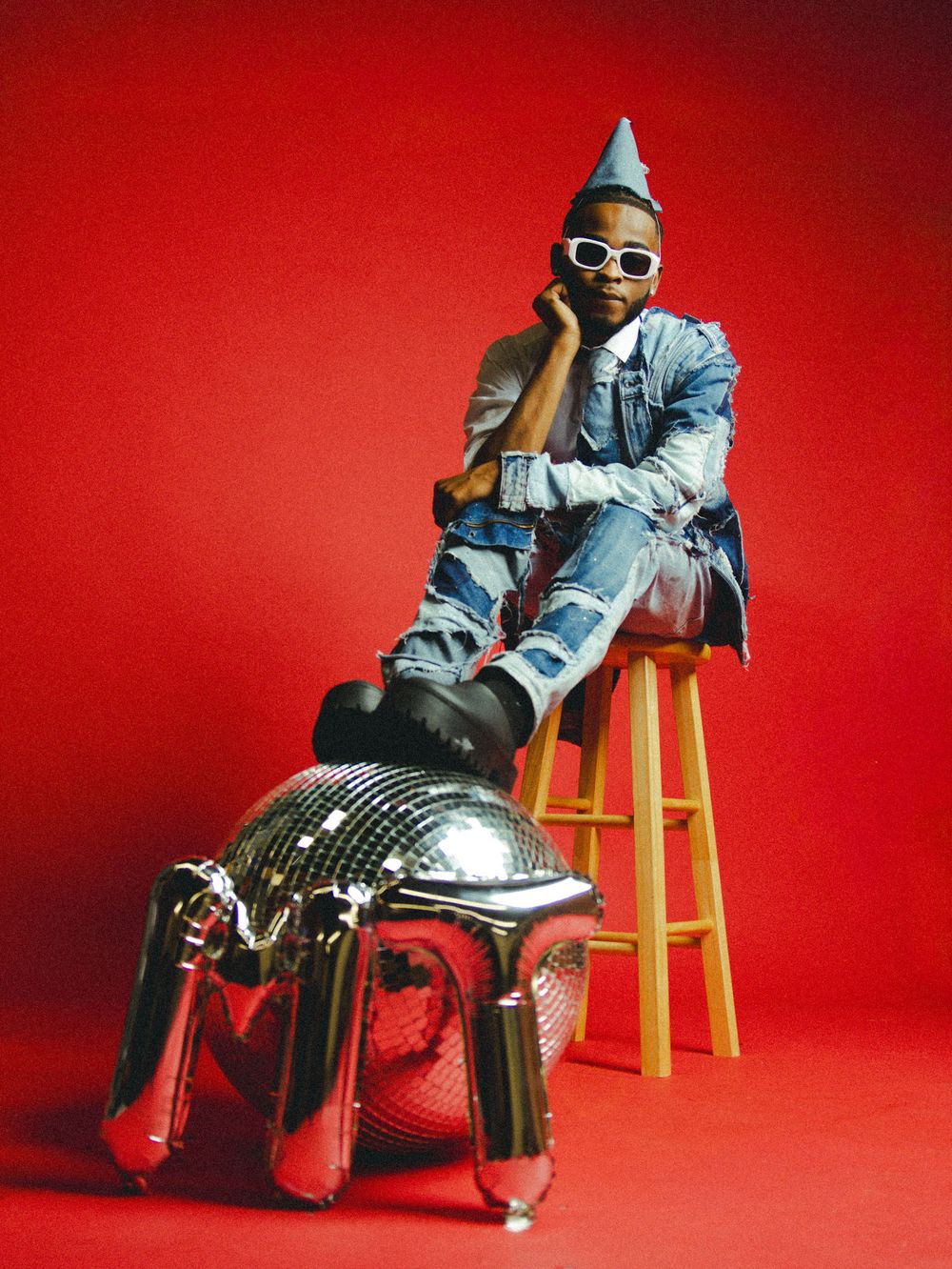 Stylish figure in denim outfit sits on yellow stool against red background.