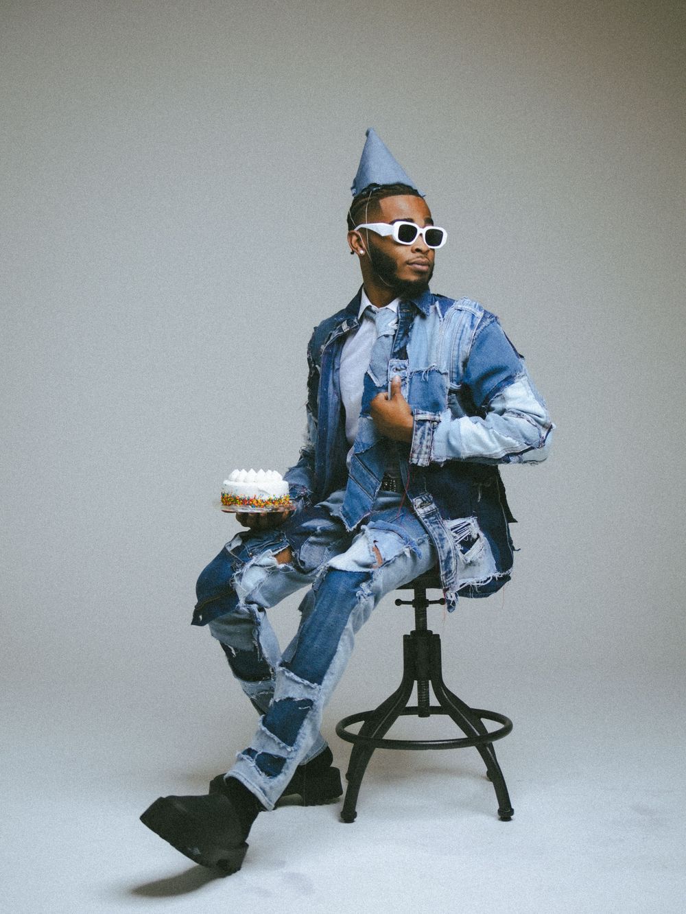 Figure in denim jacket and party hat poses playfully on studio stool.