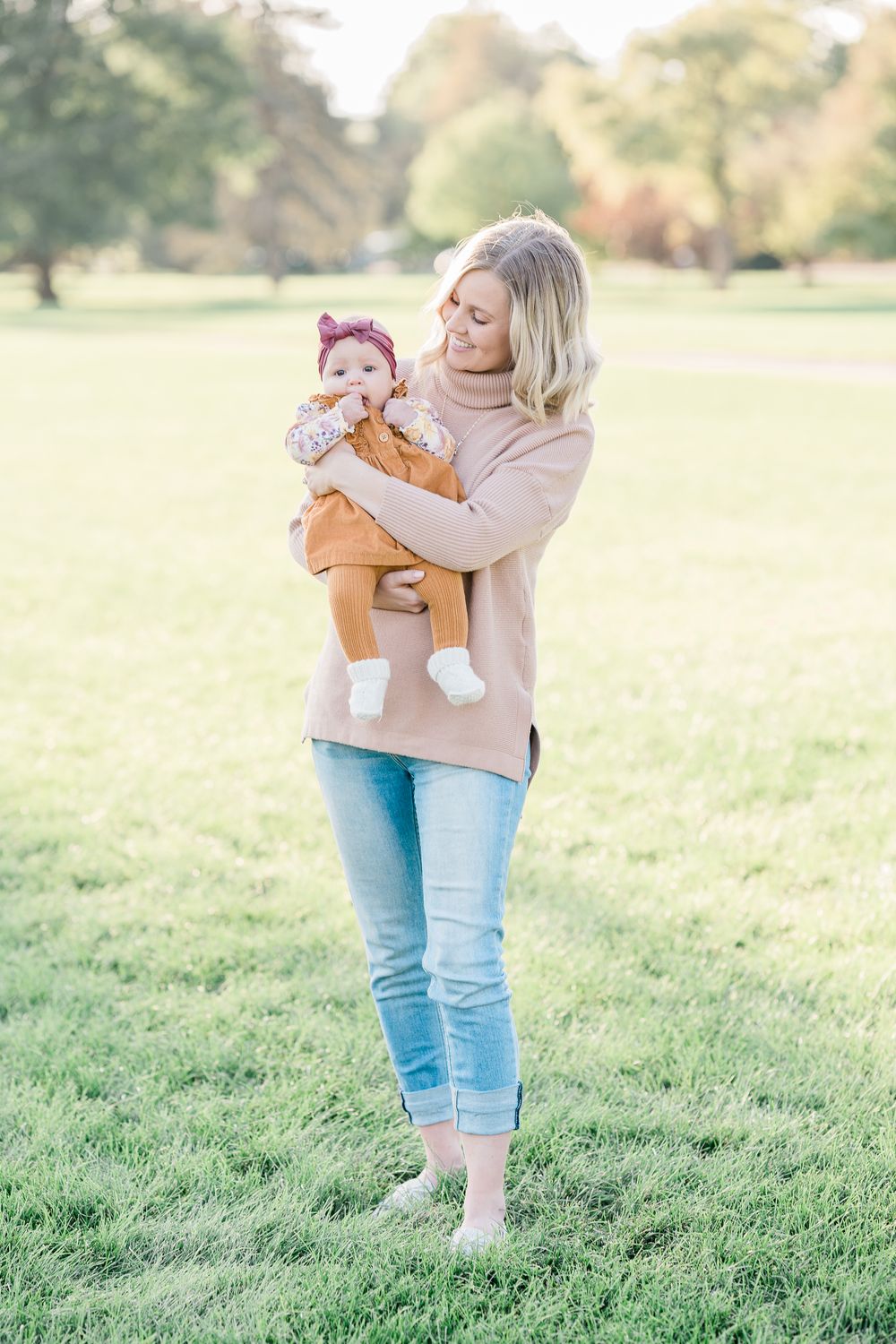 A parent wearing a beige sweater and jeans holds a baby in an orange outfit on a sunny day in a grassy field.