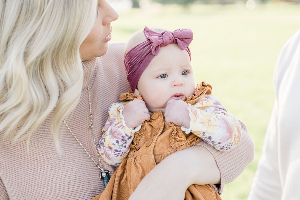 A baby wearing a brown jumper and pink headband is being held while outdoors in natural lighting.