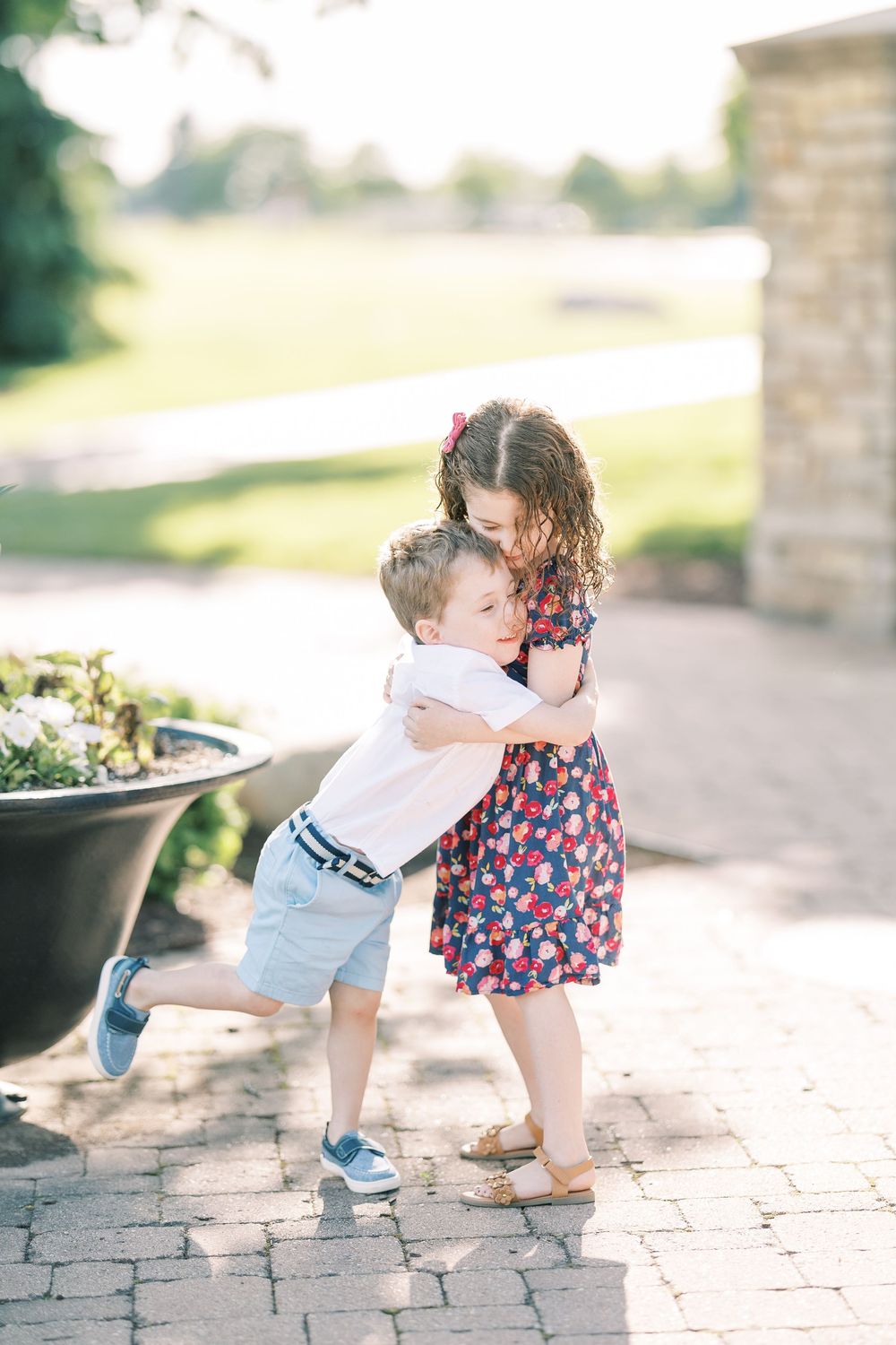Two young children sharing a sweet embrace on a brick patio near a garden planter on a sunny day.