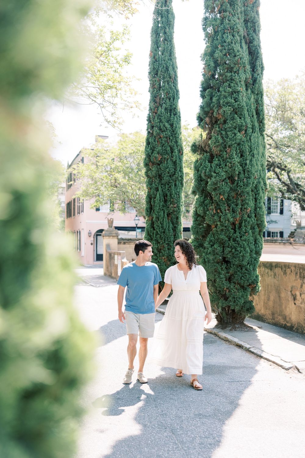 A couple walks together along a charming path lined with tall cypress trees and stone walls in a sunlit garden.