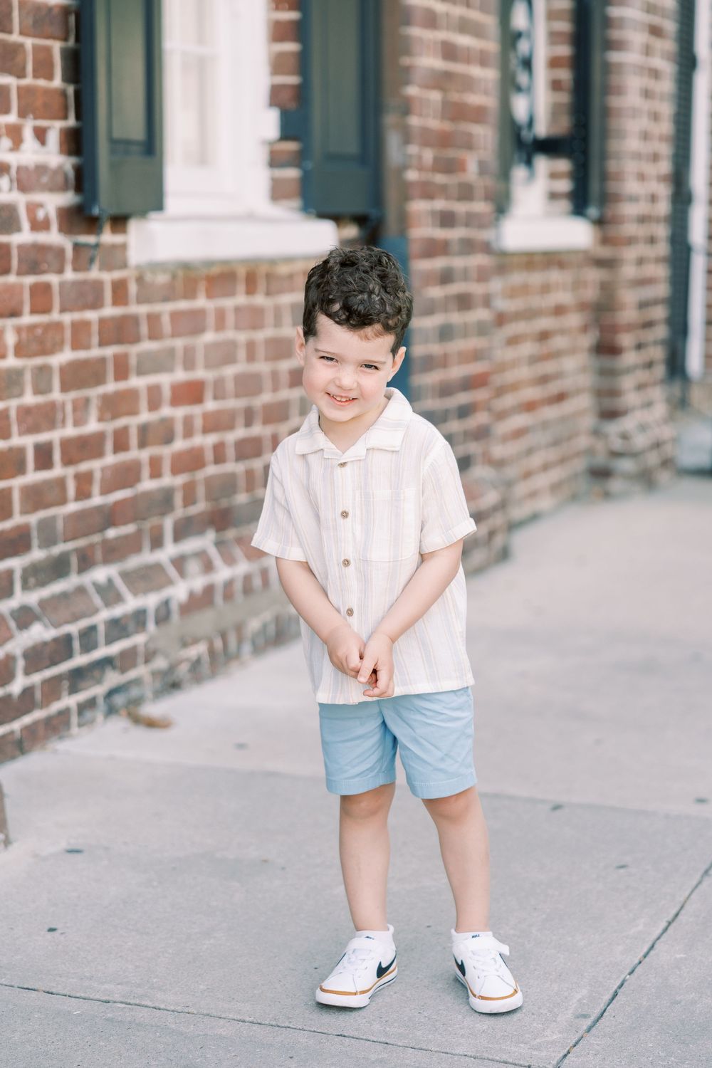 A young child stands on a sidewalk wearing a white short-sleeve shirt, blue shorts and white sneakers against a brick wall.