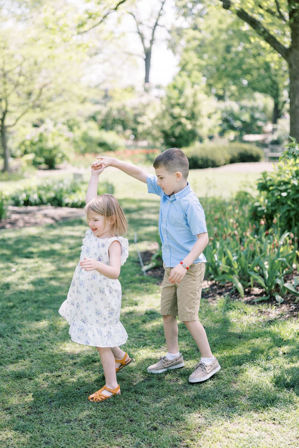 Two children dancing and twirling together in a sunny garden with blooming flowers and trees.