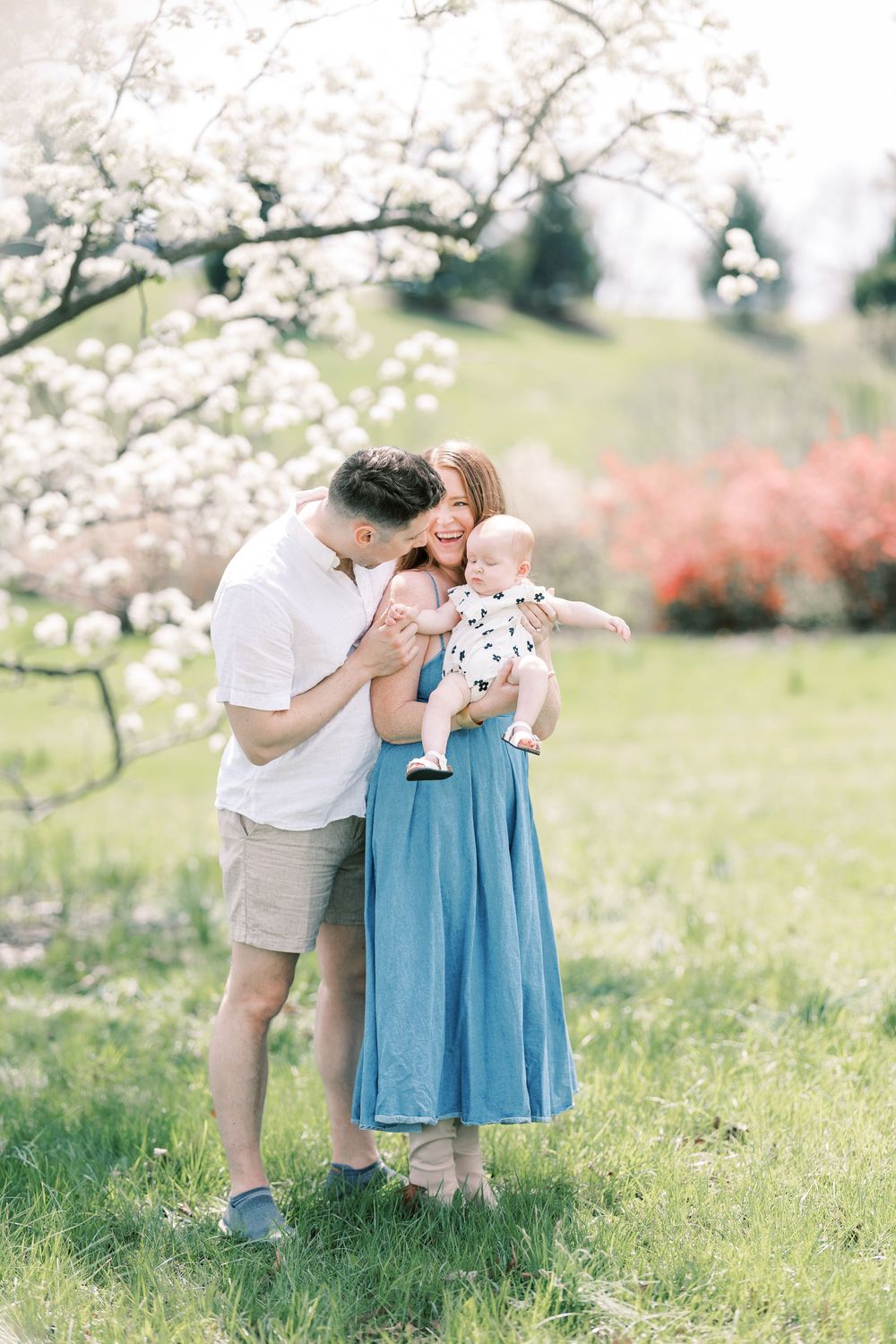 A family shares a tender moment under blooming cherry blossoms in a spring garden with white and pink flowers.