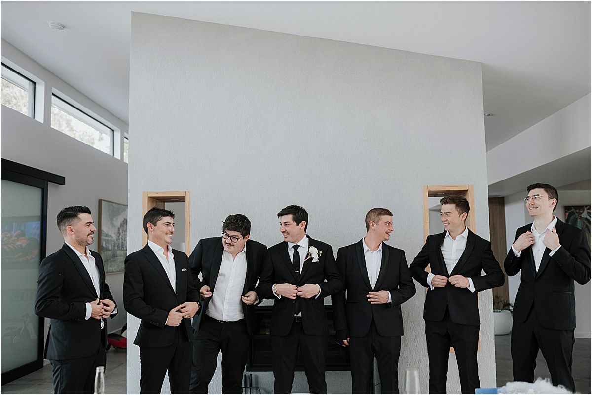 Group of groomsmen in black suits sharing a laugh in a modern white room.