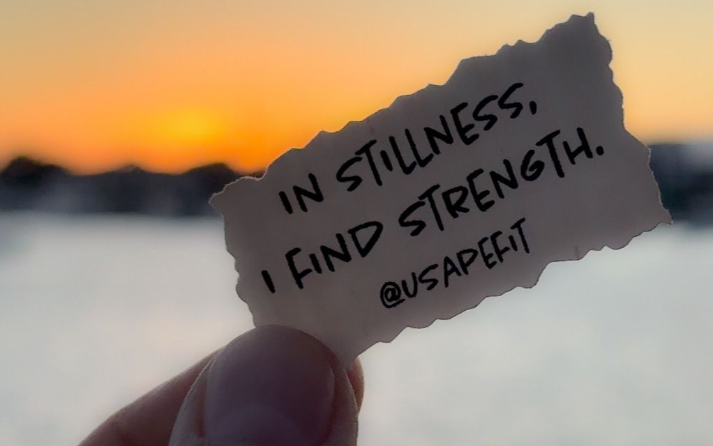 A hand holds a torn paper with the quote 'In stillness, I find strength' against a blurred sunset backdrop.