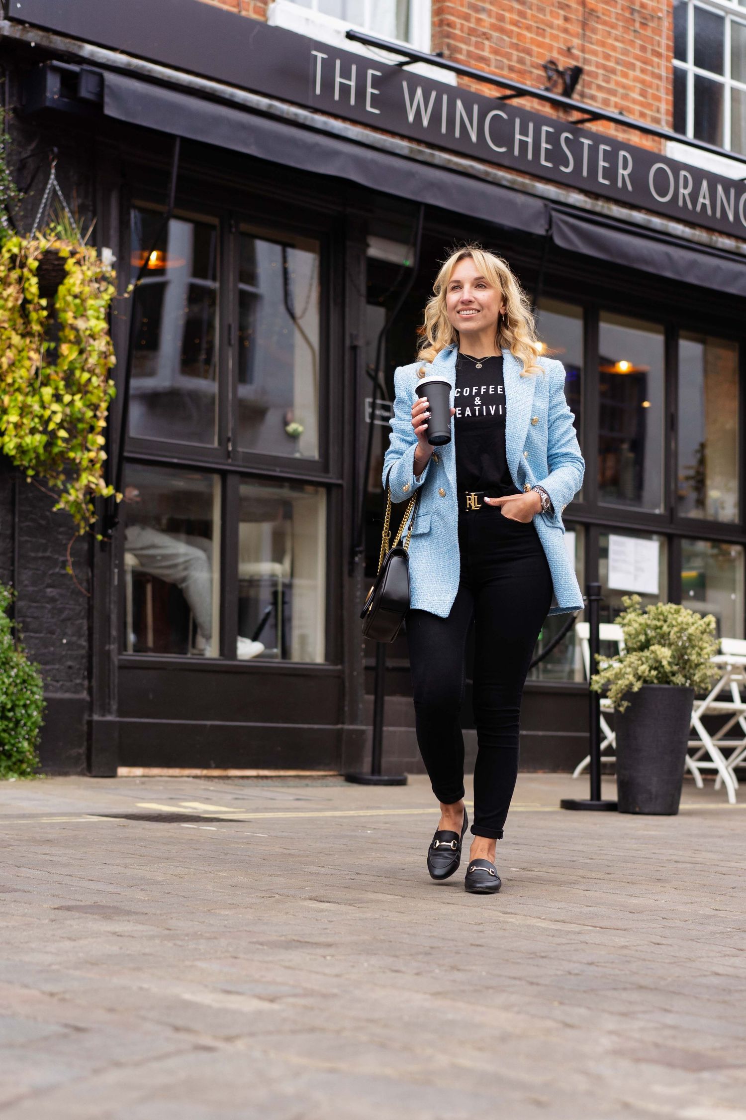 Personal branding photoshoot in Winchester shows a woman wearing a blue jacket walking casually holding a coffee outside Winchester Orangery