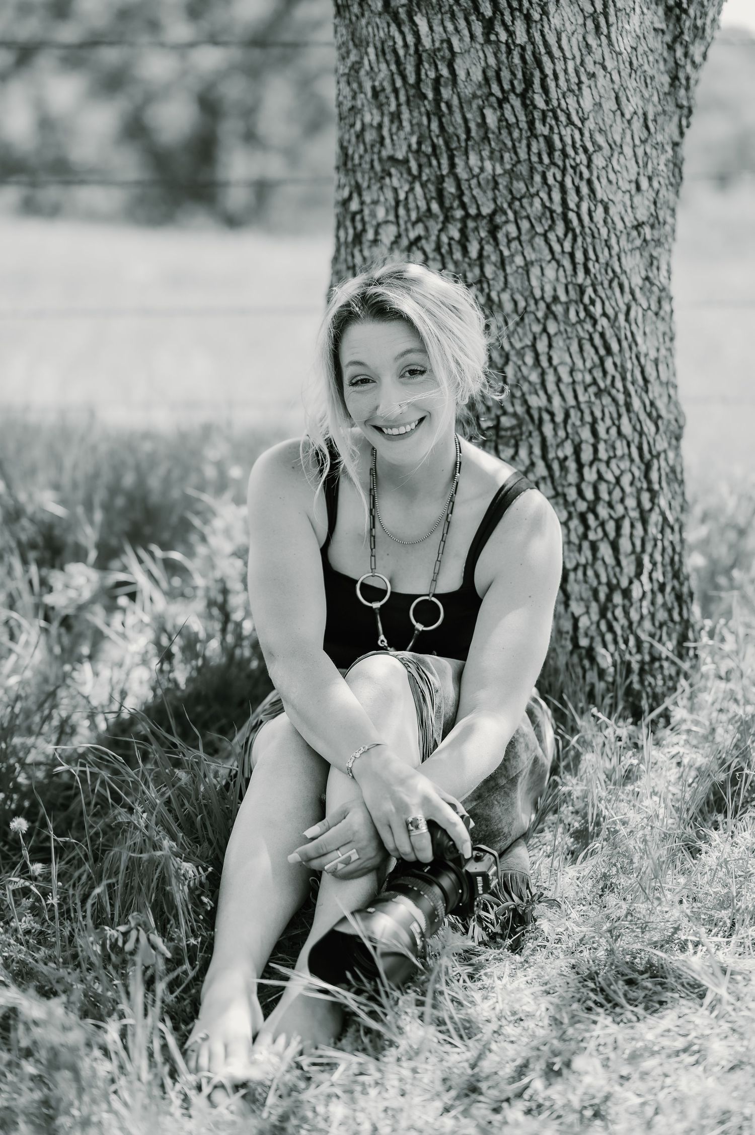 Woman in burgundy lace top with short blonde hair poses expressively against tree trunks in outdoor setting.
