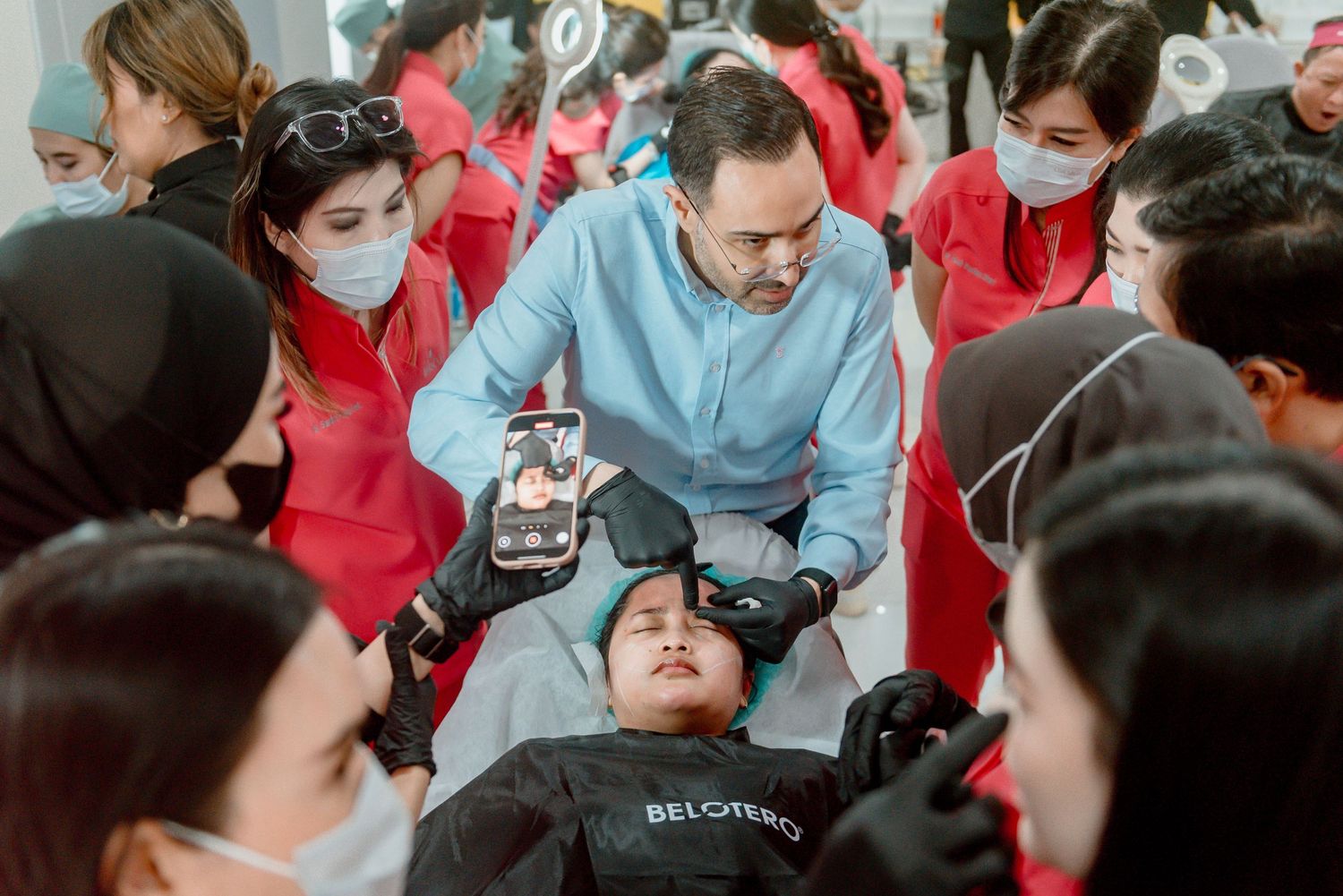 Medical students in red and blue scrubs gather around a patient simulation table during a hands-on training session.