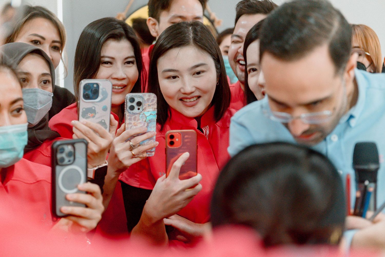 Group of people in red jackets and face masks hold up smartphones to take photos at a public event.