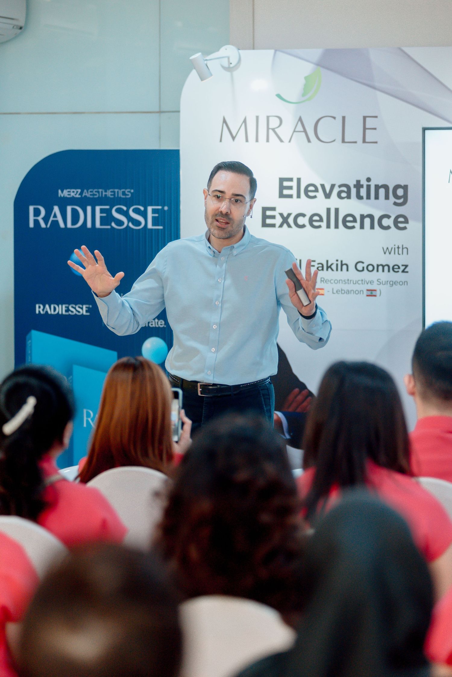 A speaker presents at a Radiesse event titled 'Elevating Excellence' to an audience wearing red shirts.