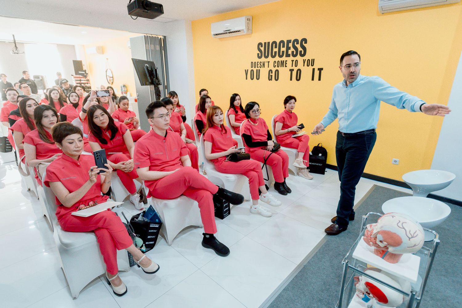 Students in matching red uniforms sit in a classroom while an instructor in blue gestures during a presentation.