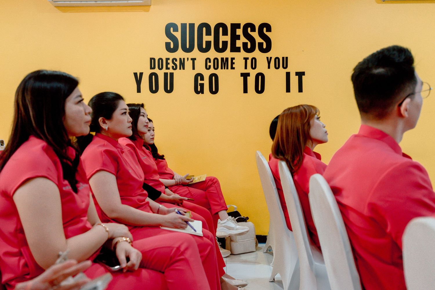 A row of people in red shirts sit at desks against a yellow wall with motivational text about success.